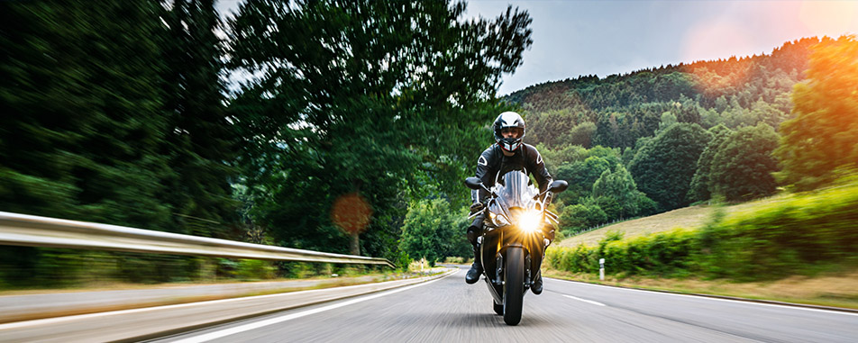 A motorcyclist wearing black gear rides swiftly down a scenic road flanked by lush trees and hills - purplecowinsurance.com