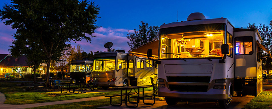 A serene RV park at dusk features multiple illuminated RVs lined up beside picnic tables - purplecowinsurance.com